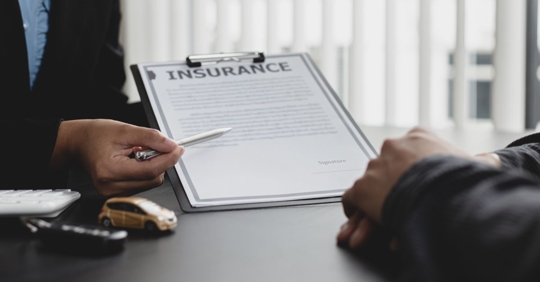 two people wearing suits sitting at a desk, one person is holding a paper on a clipboard labeled "Insurance" and pointing a pen at the page
